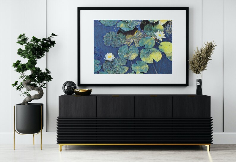 Op de afbeelding: Een zwart ingelijst beeld met een witte passe-partout, met een foto van witte waterlelies met groene bladeren in een vijver. Het beeld hangt aan een witte muur boven een zwarte kast met gouden accenten.