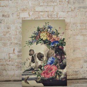 May include: A vintage still life print featuring a human skull adorned with a colorful floral arrangement. The skull is surrounded by various flowers, including red, pink, blue, and yellow blooms. The artwork is set against a light beige background.