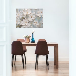 a dining area with a wooden table surrounded by modern brown chairs, adorned with a pinkish bouquet and a single white flower in a blue vase. Above the table hangs a large painting of white flowers against a muted background