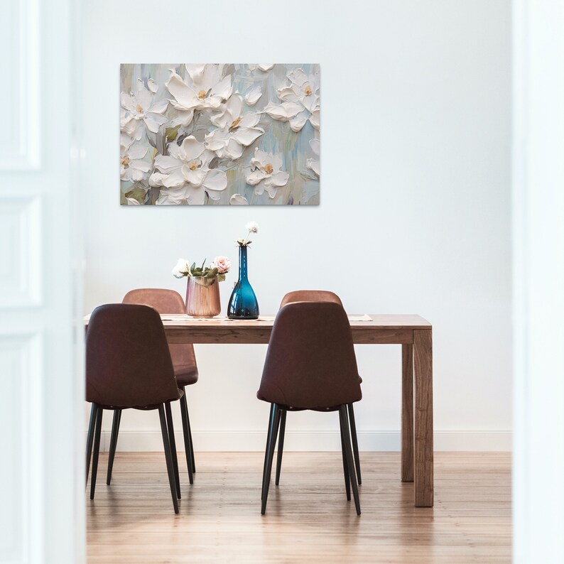 a dining area with a wooden table surrounded by modern brown chairs, adorned with a pinkish bouquet and a single white flower in a blue vase. Above the table hangs a large painting of white flowers against a muted background