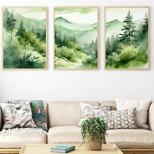 Può includere: Tre dipinti ad acquerello incorniciati che raffigurano un paesaggio boschivo verde con montagne sullo sfondo. I dipinti sono appesi a una parete bianca sopra un divano beige con cuscini e una pianta in vaso.