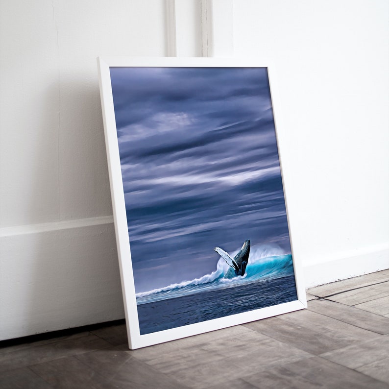 May include: A framed print featuring a breaching whale in a wave. The image shows a whale's tail emerging from a turquoise wave under a dramatic, cloudy sky. The frame is white, and the print is displayed on a wooden floor.