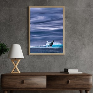 May include: A framed print featuring a whale breaching the ocean's surface. The image captures the whale's tail against a backdrop of blue water and a cloudy sky. The artwork is displayed in a wooden frame, suitable for home decor.
