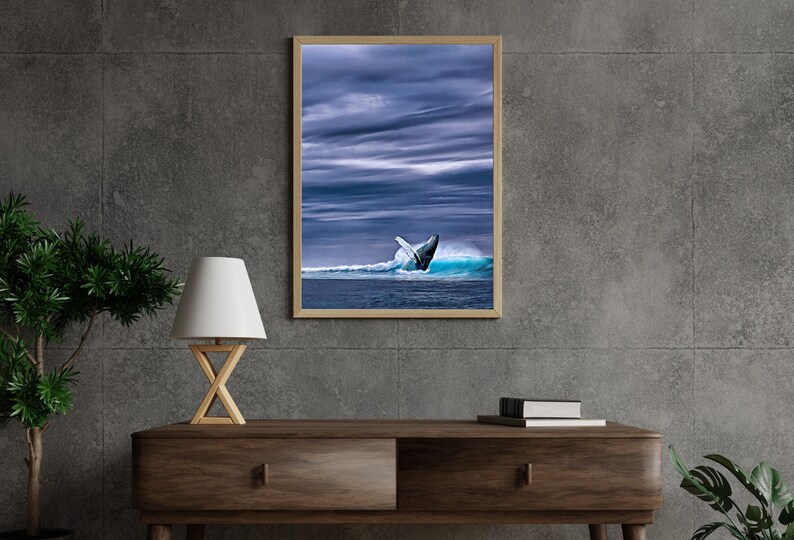 May include: A framed print featuring a whale breaching the ocean's surface. The image captures the whale's tail against a backdrop of blue water and a cloudy sky. The artwork is displayed in a wooden frame, suitable for home decor.