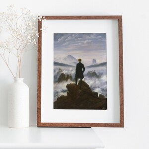 May include: A framed print of a painting depicting a man standing on a mountaintop overlooking a sea of clouds. The man is wearing a dark coat and is holding a walking stick. The sky is a light blue with some clouds.