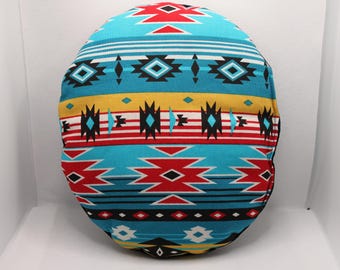 May include: A round throw pillow featuring a vibrant geometric pattern in turquoise, red, yellow, white, and black. The design consists of horizontal bands with repeating diamond, chevron, and striped motifs across the entire surface of the cushion.