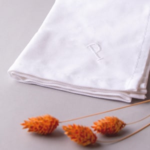 May include: A white linen handkerchief with a single embroidered letter "P" in the center. The handkerchief is folded and lying on a gray surface with orange dried flowers in the foreground.
