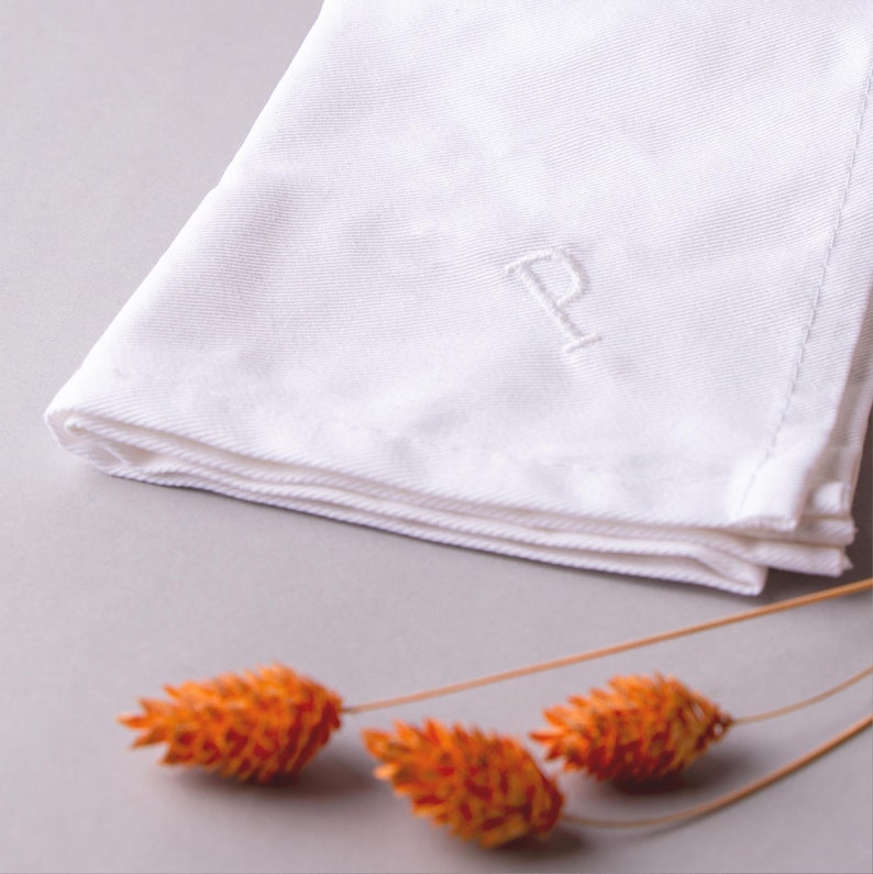 May include: A white linen handkerchief with a single embroidered letter "P" in the center. The handkerchief is folded and lying on a gray surface with orange dried flowers in the foreground.