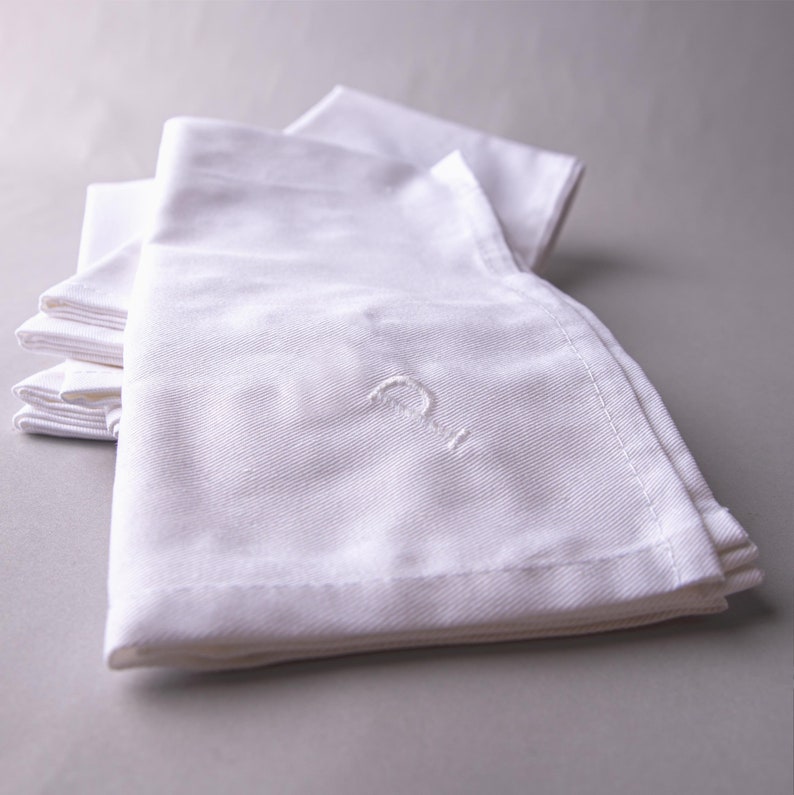 May include: A stack of white linen napkins with a single initial embroidered on the top napkin.