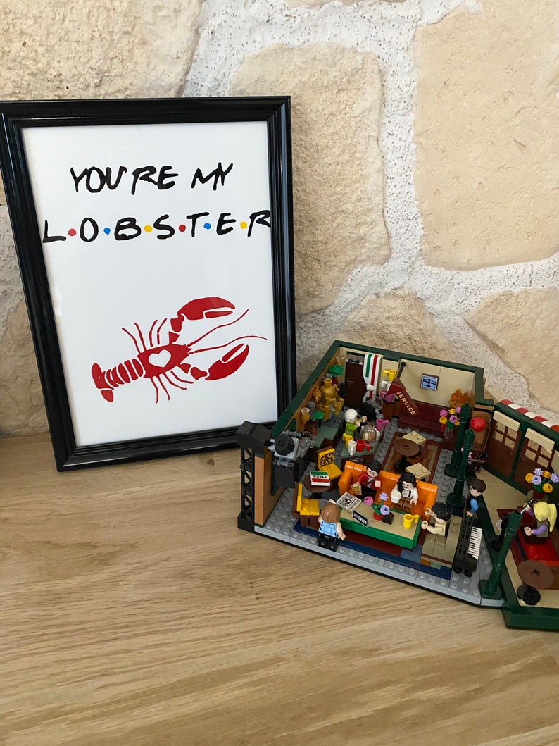 May include: A framed print with the text "YOU'RE MY L.O.B.S.T.E.R" and a red lobster illustration. Beside it is a Lego set depicting a cafe scene with miniature figures and furniture.