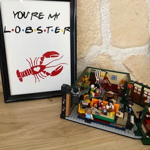 May include: A framed print with the text "YOU'RE MY L.O.B.S.T.E.R" and a red lobster illustration. Beside it is a detailed LEGO model of a cafe interior, featuring miniature figures and furniture.