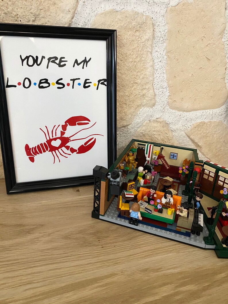 May include: A framed print with the text "YOU'RE MY L.O.B.S.T.E.R" and a red lobster illustration. Beside it is a detailed LEGO model of a cafe interior, featuring miniature figures and furniture.