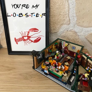 May include: A framed print with the text "YOU'RE MY L.O.B.S.T.E.R" and a red lobster illustration. Beside it is a detailed LEGO model of a cafe interior, featuring miniature figures and furniture.