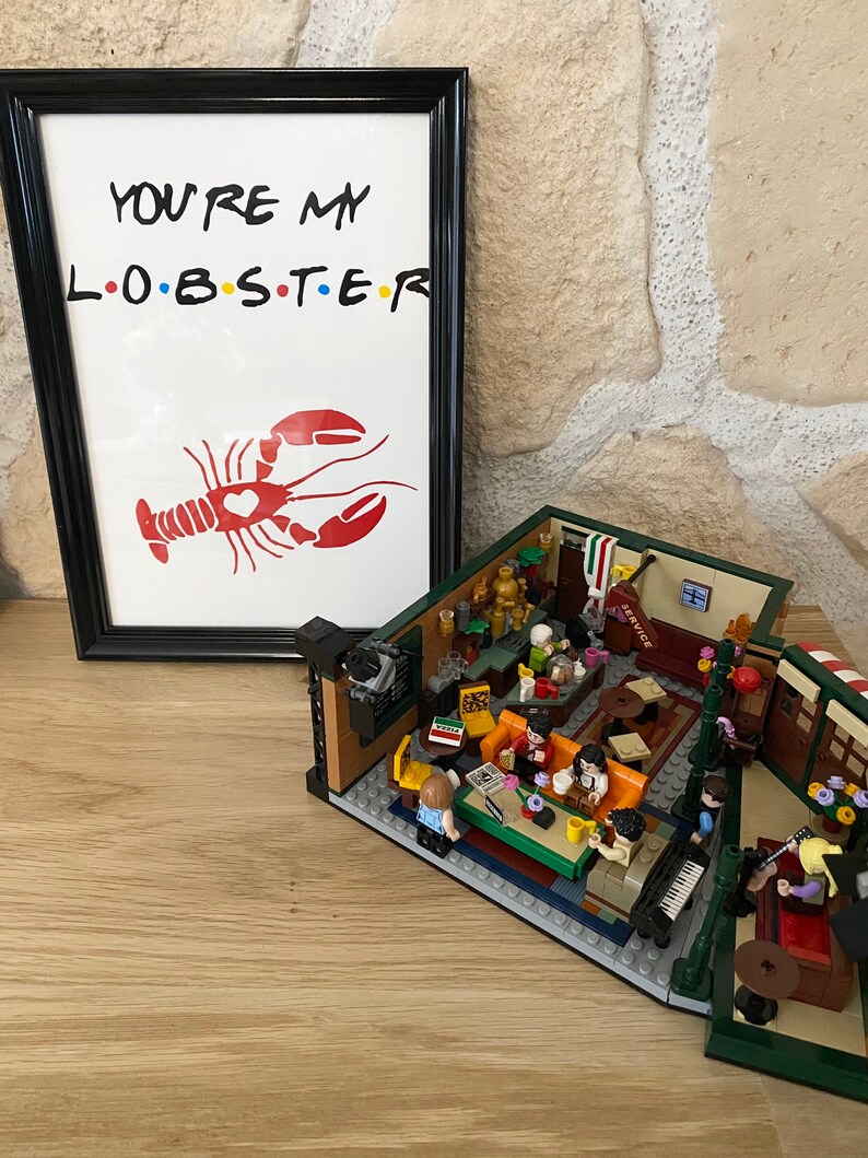May include: A framed print with the text "YOU'RE MY L.O.B.S.T.E.R" and a red lobster illustration. Beside it is a detailed LEGO model of a cafe interior, featuring miniature figures and furniture.