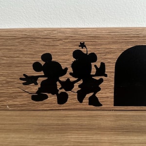 Peut inclure: Silhouette noire de Mickey Mouse et Minnie Mouse se tenant la main, avec une silhouette noire d'un trou de souris à droite. Les silhouettes sont sur une surface en bois brun clair.