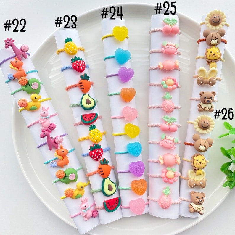 May include: A set of 10 colorful hair ties with various charms, including hearts, fruit, animals, and candy. The hair ties are numbered 22 through 26.