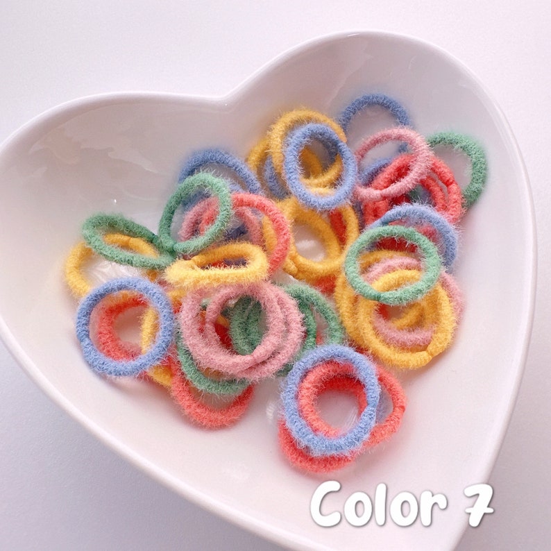 May include: A bowl of colorful fuzzy hair ties in blue, green, yellow, and pink. The bowl is heart-shaped and white. The text "Color 7" is written below the bowl.