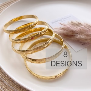 May include: A stack of five gold-toned metal bangles, each featuring a textured surface with a repeating geometric pattern. The bracelets are arranged in a cascading formation. A small card with the text LittleHoop Treasures and 8 DESIGNS is visible.
