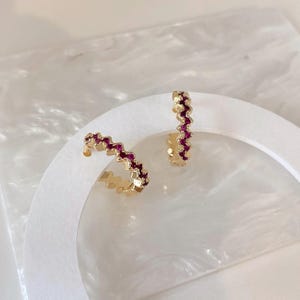 Könnte beinhalten: Ein Paar goldene Creolen mit einem Zickzack- oder Chevron-Design. Jeder Ohrring ist mit einer Reihe kleiner, leuchtend rosaroter Edelsteine akzentuiert, die entlang der Außenkante der goldenen Metallstruktur eingebettet sind.