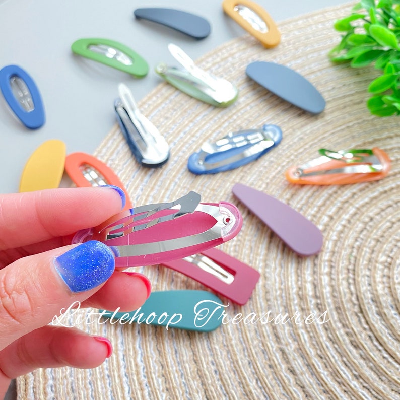Puede incluir: Primer plano de una pinza para el pelo de color rosa con un cierre de metal plateado. La pinza se sostiene en una mano con esmalte de u&ntilde;as azul con purpurina. La pinza est&aacute; rodeada de otras pinzas para el pelo de varios colores, entre ellos amarillo, naranja, azul, verde y morado.