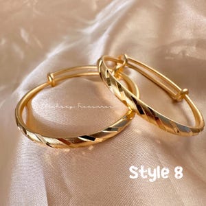 May include: Two gold-colored adjustable bangles with a textured, geometric design. The bangles are displayed on a satin surface. The text "Style 8" is visible in the image.