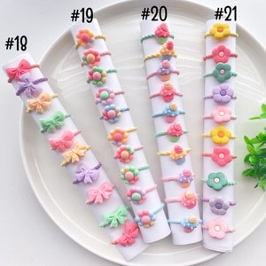 May include: A set of 10 colorful hair ties with different designs, including bows, flowers, and cupcakes. The hair ties are arranged in rows on a white plate, labeled with numbers 18 through 21.