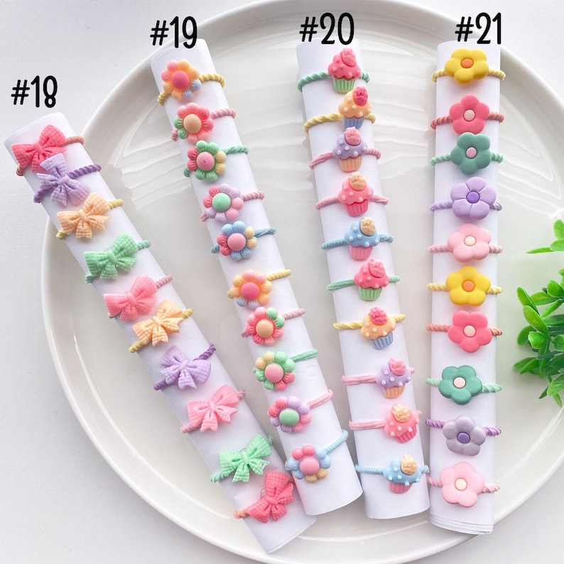 May include: A set of 10 colorful hair ties with different designs, including bows, flowers, and cupcakes. The hair ties are arranged in rows on a white plate, labeled with numbers 18 through 21.