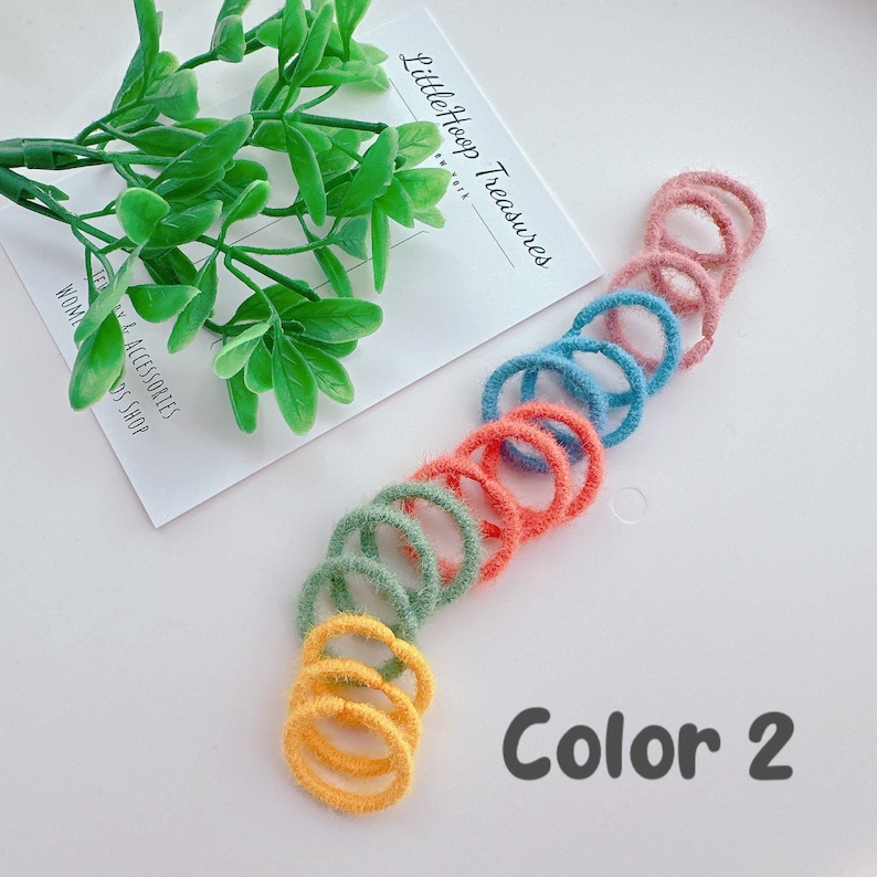 Puede incluir: Un conjunto de 10 gomas para el pelo de color borroso en tonos de verde, azul, naranja y amarillo. Las gomas para el pelo est&aacute;n dispuestas en semic&iacute;rculo sobre una superficie blanca. El texto "Color 2" est&aacute; escrito en negro debajo de las gomas para el pelo.