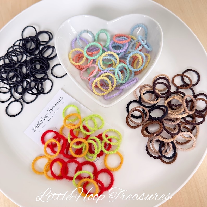 May include: Assortment of colorful hair ties in a variety of colors, including black, red, yellow, green, light blue, pink, brown, and beige. The hair ties are arranged in four piles on a white plate. The text "Little Hoop Treasures" is visible on a white card.