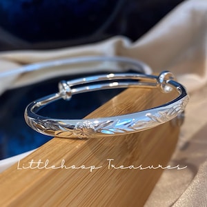 Puede incluir: Una pulsera tipo brazalete extensible de tono plateado con acabado pulido y patrones de hojas grabados en la banda exterior. La pulsera tiene dos conectores deslizantes circulares que permiten ajustar el tamaño. El texto Littlehoop Treasures es visible.