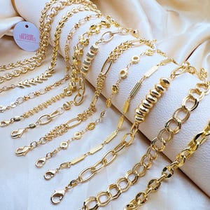 Peut inclure: Un ensemble de 12 bracelets en chaîne dorée avec différents styles et tailles de maillons. Les bracelets sont disposés sur un fond en tissu blanc doux. Le texte "Little Shop Treasures" est visible sur une étiquette blanche.