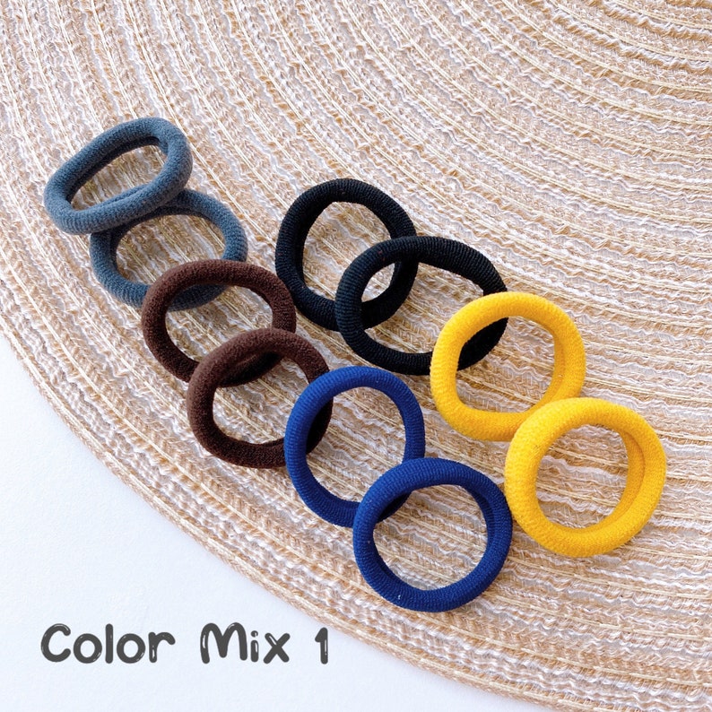 May include: A set of eight hair ties in various colors, including navy blue, dark brown, black, and yellow. The hair ties are arranged in a circle on a woven surface. The text "Color Mix 1" is visible below the hair ties.