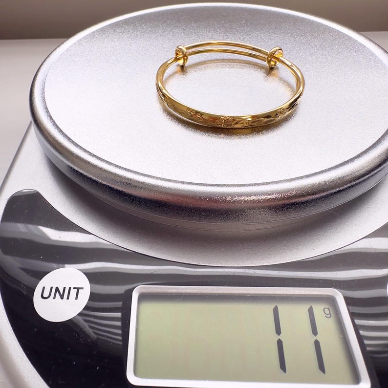 May include: A gold-colored adjustable bangle bracelet with etched details, resting on a silver scale. The scale displays a weight of 11 grams. The bracelet is a piece of jewelry.