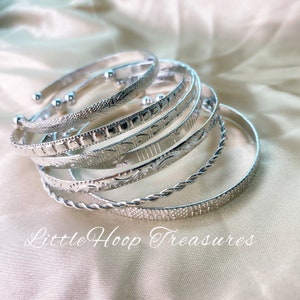May include: A set of seven silver bangles with different engraved patterns. The bangles are stacked on top of each other and have a shiny finish. The text "Little Hoop Treasures" is visible at the bottom of the image.