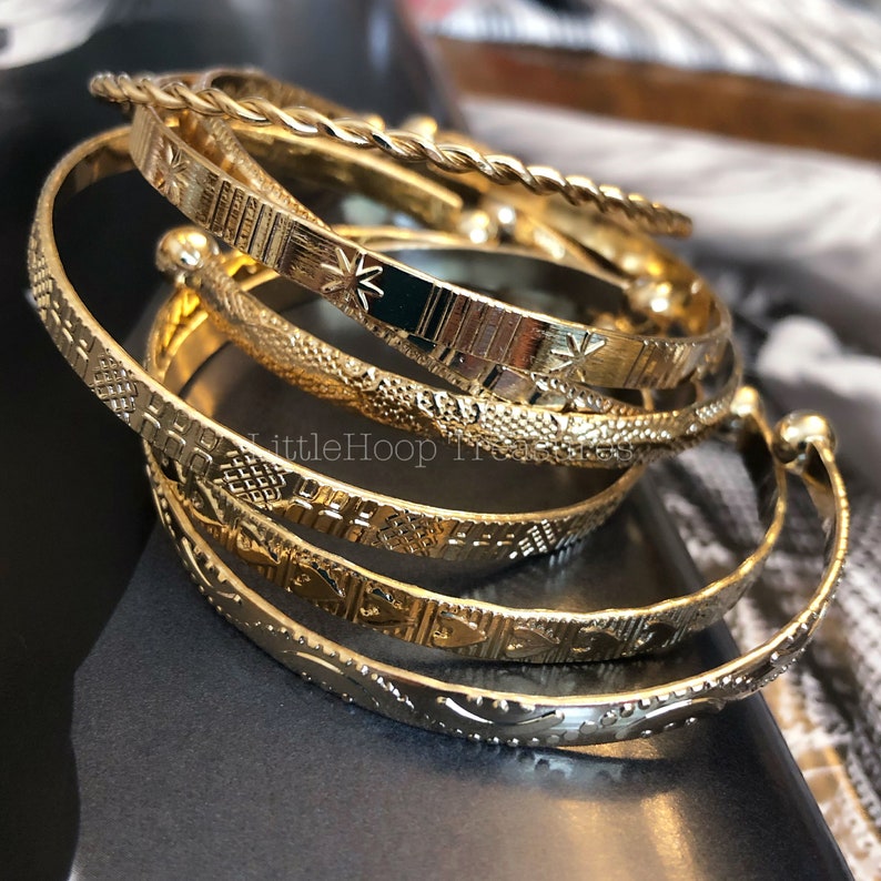 May include: A set of five gold-toned bangle bracelets with intricate patterned designs. The bracelets are stacked on top of each other, creating a layered look.