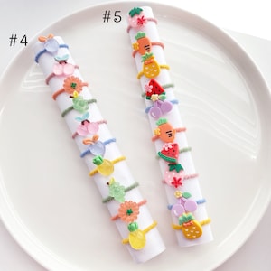 May include: Two sets of colorful hair ties with various fruit and flower charms. The hair ties are arranged in a vertical line on a white plate. The text #4 and #5 are visible on the plate.