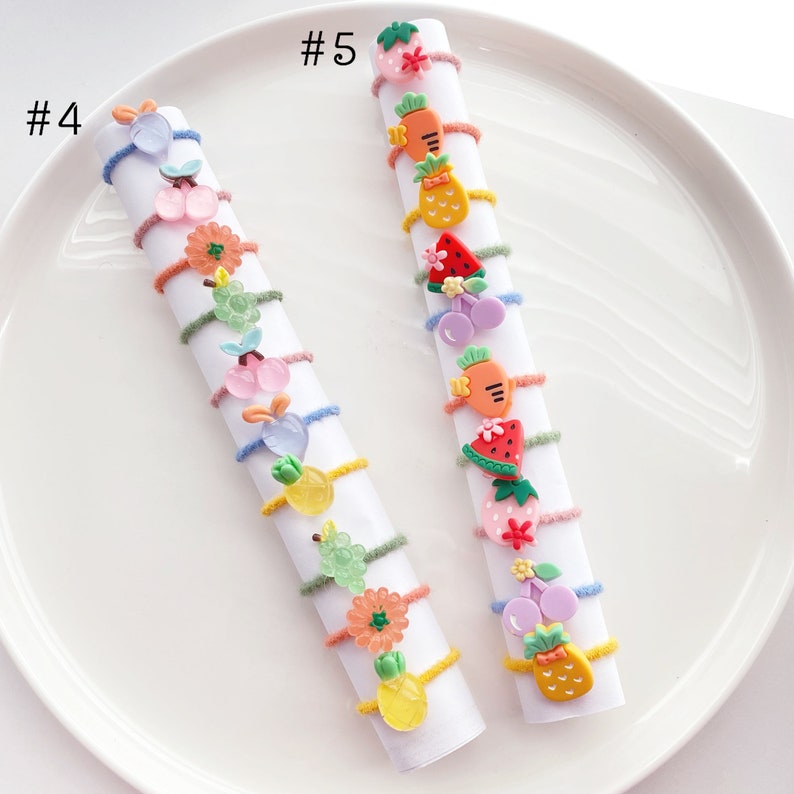 May include: Two sets of colorful hair ties with various fruit and flower charms. The hair ties are arranged in a vertical line on a white plate. The text #4 and #5 are visible on the plate.