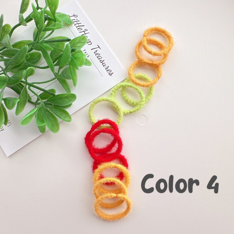 Puede incluir: Un conjunto de gomas para el pelo de colores rojo, amarillo y verde. Las gomas para el pelo est&aacute;n hechas de material peludo y est&aacute;n apiladas. El texto "Color 4" est&aacute; escrito en negro sobre un fondo blanco.