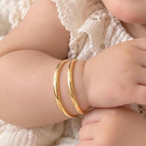 May include: Two polished gold bangles with a smooth, rounded surface. These circular bracelets feature a reflective metallic finish and a simple, continuous band design, worn together on a wrist to create a layered appearance.