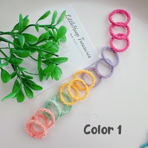 Puede incluir: Un conjunto de 7 scrunchies peludos de colores brillantes en un esquema de colores arco&iacute;ris. Los scrunchies est&aacute;n dispuestos en l&iacute;nea sobre una superficie blanca. El texto "Little Hoop Treasures" y "Color 1" es visible en la imagen.