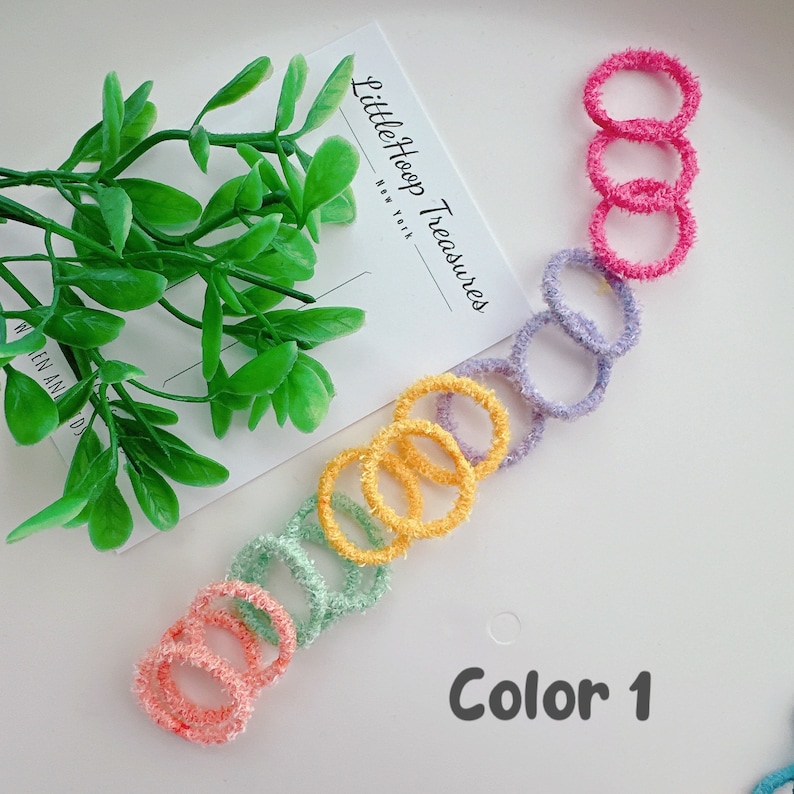 Puede incluir: Un conjunto de 7 scrunchies peludos de colores brillantes en un esquema de colores arco&iacute;ris. Los scrunchies est&aacute;n dispuestos en l&iacute;nea sobre una superficie blanca. El texto "Little Hoop Treasures" y "Color 1" es visible en la imagen.