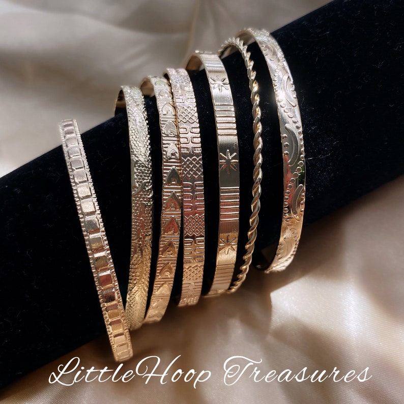 May include: A set of six gold-toned bangle bracelets with different engraved patterns. The bracelets are stacked on top of each other on a black surface.  Little Hoop Treasures.