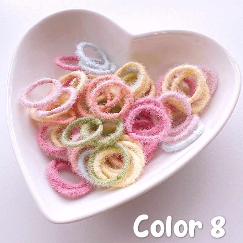 May include: A heart-shaped bowl filled with a variety of colorful fuzzy hair ties. The hair ties are in shades of pink, yellow, green, and blue. The bowl is on a white surface. Color 8.
