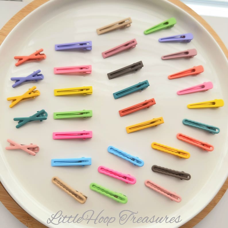 May include: A collection of 20 colorful plastic hair clips in various shapes and sizes. The clips are arranged on a white plate. The clips are in shades of pink, blue, green, yellow, orange, and brown.
