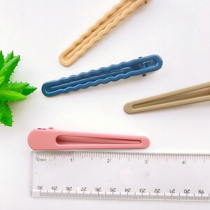 Op de afbeelding: Vier haarclips in verschillende kleuren: beige, blauw, bruin en roze. De clips zijn allemaal ongeveer 7,5 cm lang en hebben een golvend ontwerp. Een liniaal bevindt zich onderaan de afbeelding.
