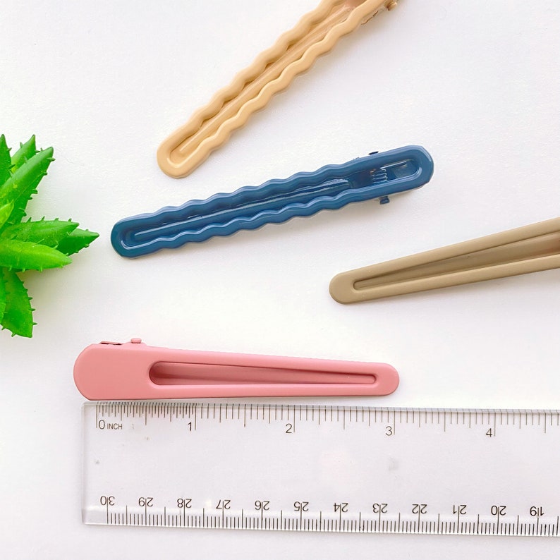 Op de afbeelding: Vier haarclips in verschillende kleuren: beige, blauw, bruin en roze. De clips zijn allemaal ongeveer 7,5 cm lang en hebben een golvend ontwerp. Een liniaal bevindt zich onderaan de afbeelding.