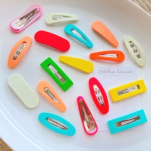 Op de afbeelding: Assortiment van kleurrijke plastic haarclips in verschillende vormen en maten. De clips zijn gerangschikt op een wit bord. De tekst "Little Hoop Treasures" is zichtbaar op een witte kaart.