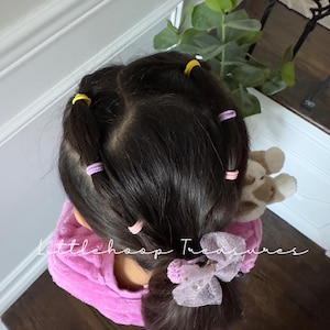 May include: A young person with long dark hair styled with multiple pink and yellow hair ties. The hair is pulled back into a ponytail with a sparkly pink bow.