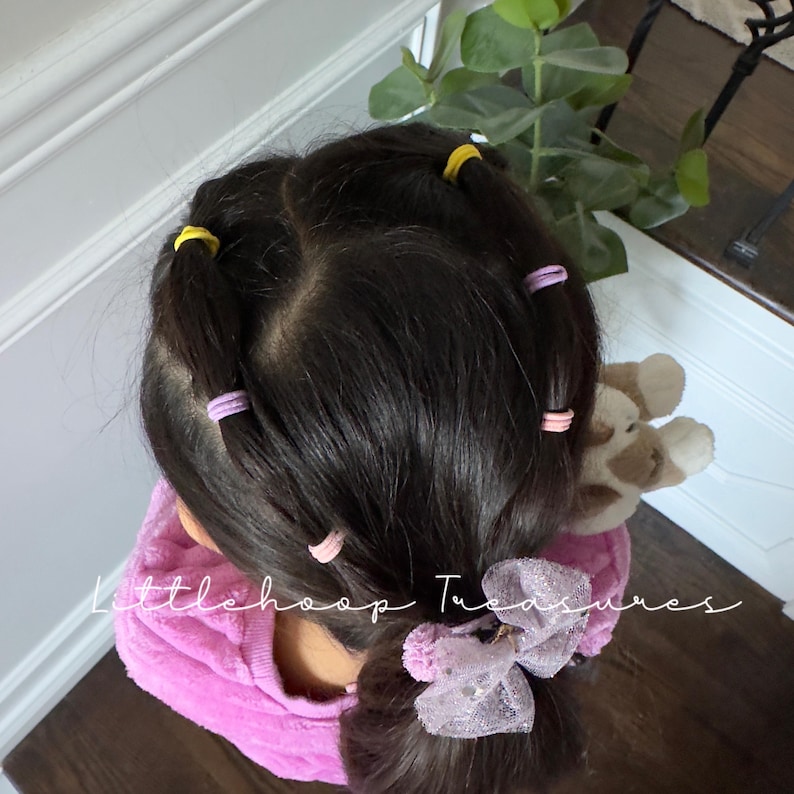 May include: A young person with long dark hair styled with multiple pink and yellow hair ties. The hair is pulled back into a ponytail with a sparkly pink bow.