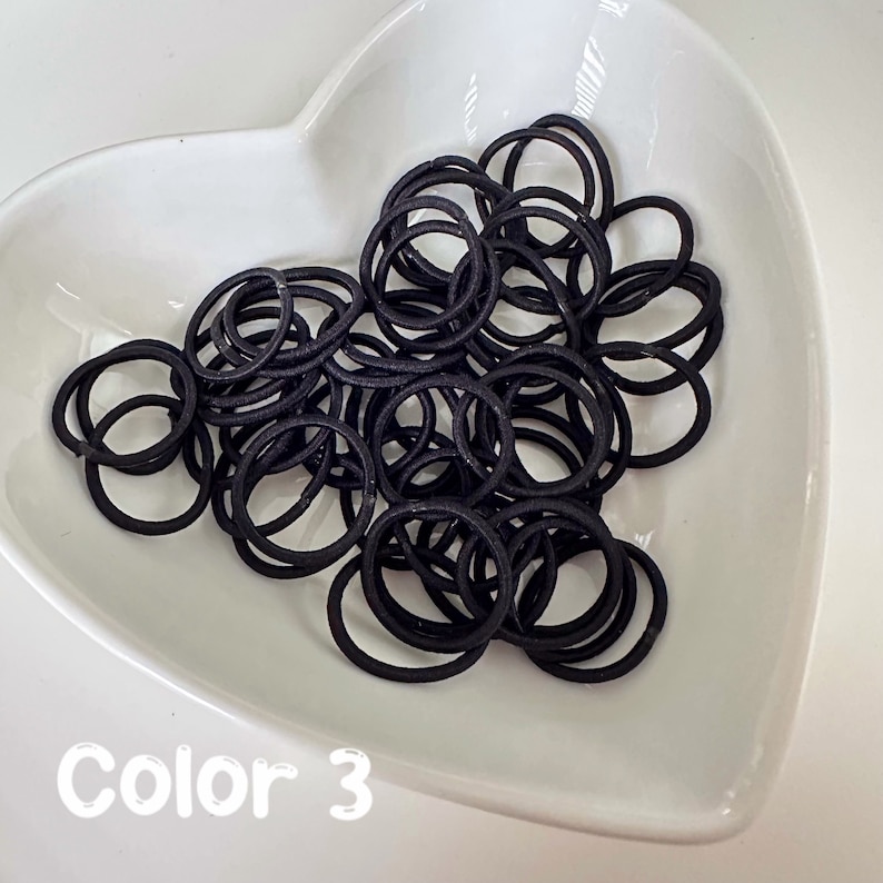 May include: A white heart-shaped dish filled with a large quantity of black hair elastics. The text "Color 3" is written below the dish.